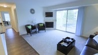 This single-floor living space offers a well-appointed layout centered around a stylish corner fireplace framed in light stone or tile, which serves as a natural focal point for the room. A wall-mounted flat-screen television is positioned above the fireplace, creating a practical and visually cohesive entertainment area. The living area features a combination of hardwood-style flooring and a plush area rug, adding warmth and texture underfoot. Dark leather accent chairs with green throw pillows provide comfortable seating, complementing the room's neutral color palette of soft whites and grays. A pair of modular ottoman-style coffee tables in dark wood offer both functionality and a modern aesthetic. One of the most notable features of this space is the expansive sliding glass door that floods the room with natural light and opens onto a private balcony, offering pleasant outdoor views of a neighboring building and greenery. The walls are clean and minimal, highlighted by a decorative sunburst mirror and a slender floral arrangement. The overall ambiance is bright, open, and inviting, with a balanced mix of modern and cozy design elements throughout.