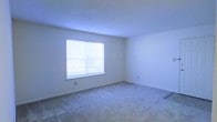 This single-floor residential space offers a straightforward and functional layout designed for comfortable everyday living. The interior is characterized by neutral-toned walls and smooth ceilings with a subtle texture, creating a clean and understated aesthetic throughout. The primary room is generously proportioned, featuring wall-to-wall carpeting in a light, neutral shade that adds warmth and comfort underfoot. A centrally positioned window, outfitted with horizontal blinds, allows natural light to filter in and illuminate the space, contributing to an open and airy atmosphere. The room's simplicity provides a versatile canvas well-suited to a variety of furnishing arrangements and personal styles. Electrical outlets are conveniently placed along the baseboards, ensuring practical usability throughout. The overall condition of the space appears well-maintained and move-in ready, with clean surfaces and an uncluttered environment. The design prioritizes functionality while maintaining a bright and welcoming ambiance, making it an accessible and adaptable living environment suitable for a range of occupants.