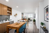 This single-floor residence showcases a thoughtfully designed open-concept layout that seamlessly connects the kitchen, dining, and living areas. The kitchen is equipped with warm wood cabinetry, a gray subway tile backsplash, white countertops, and a full suite of stainless steel appliances, including a range, microwave, dishwasher, and refrigerator. A butcher-block dining table with teal upholstered counter stools adds a functional yet stylish centerpiece to the space. Light hardwood flooring runs throughout, enhancing the bright and airy atmosphere created by recessed lighting. The living area is anchored by floor-to-ceiling sliding glass doors that invite natural light and offer views of the surrounding outdoor courtyard. The bedroom also features large windows with outdoor views, providing a calm, light-filled retreat. A walk-in closet area is accessible off the bedroom. The bathroom includes a full soaking tub and shower combination with clean white tile surround. Throughout the residence, crisp white walls serve as a backdrop for vibrant art and mid-century modern–inspired furnishings, creating a cohesive, contemporary aesthetic that balances warmth and style.