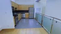 This single-floor space offers a thoughtfully designed layout with a fully equipped kitchen as a central feature. The kitchen is outfitted with warm honey-toned wood cabinetry providing ample upper and lower storage, complemented by dark countertops that create a striking contrast. Stainless steel appliances — including a refrigerator, dishwasher, and range — add a modern, functional touch. A sink is conveniently positioned along the cabinetry run, and a large ventilation duct overhead speaks to the space's practical, utilitarian character. The kitchen area is finished with large-format gray tile flooring, which transitions seamlessly into warm hardwood flooring in the adjacent area, delineating the spaces naturally. A standout architectural detail is the sleek frosted glass panel railing with polished metal hardware, which borders the open area and adds a contemporary, refined element to the overall design. The high ceiling height and open layout lend the space an airy, expansive feel, while exposed ductwork hints at an industrial-inspired aesthetic. This space combines practicality with modern design sensibilities, making it both functional and visually engaging.
