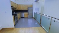 This single-floor space offers a thoughtfully designed layout with a fully equipped kitchen as a central feature. The kitchen is outfitted with warm honey-toned wood cabinetry providing ample upper and lower storage, complemented by dark countertops that create a striking contrast. Stainless steel appliances — including a refrigerator, dishwasher, and range — add a modern, functional touch. A sink is conveniently positioned along the cabinetry run, and a large ventilation duct overhead speaks to the space's practical, utilitarian character. The kitchen area is finished with large-format gray tile flooring, which transitions seamlessly into warm hardwood flooring in the adjacent area, delineating the spaces naturally. A standout architectural detail is the sleek frosted glass panel railing with polished metal hardware, which borders the open area and adds a contemporary, refined element to the overall design. The high ceiling height and open layout lend the space an airy, expansive feel, while exposed ductwork hints at an industrial-inspired aesthetic. This space combines practicality with modern design sensibilities, making it both functional and visually engaging.
