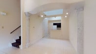 This two-floor space offers a thoughtfully designed interior that blends classic architectural character with a clean, open layout. The main living area is defined by a striking decorative stone column and a sweeping arched entryway, lending a sense of elegance and structure to the space. Light-toned ceramic tile flooring extends throughout, contributing to a bright and cohesive aesthetic.

A central skylight is recessed into a raised ceiling section, drawing natural light deep into the interior and highlighting the varying ceiling heights that add dimension to the space. Recessed and track lighting complement the natural illumination, ensuring the area remains well-lit throughout the day.

The living area flows seamlessly into the kitchen, which features dark cabinetry contrasted against light countertops and a pass-through or breakfast bar, creating an accessible and functional connection between the two spaces. A white refrigerator and built-in cabinetry are visible, offering practical storage and appliance integration.

The overall ambiance of the space is open and airy, with neutral walls and pale finishes that maximize the sense of light and space across both floors. The combination of classical design elements and modern layout makes this a versatile and visually engaging property.