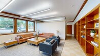 Located at 6 Delprete Avenue in Rockland, MA, this single-floor amenity space is thoughtfully designed to offer both relaxation and fitness in one compact layout. The space is divided into two distinct areas: a living room and an exercise room, each with its own character and purpose.

The living room exudes a warm, welcoming ambiance, highlighted by rich warm-toned wood wainscoting, built-in shelving units with cabinetry, and decorative crown molding. Linear recessed lighting illuminates the space evenly from above, complementing the natural light that pours in through a set of large, wood-framed windows offering views of the surrounding neighborhood. The seating area features an orange upholstered sofa, two patterned blue accent chairs, and a low wooden coffee table arranged over a neutral area rug, creating an inviting, communal atmosphere. A wall-mounted mini-split unit ensures year-round comfort.

The exercise room is energetic and functional, anchored by a vibrant Boston skyline mural that spans one full wall. The room is outfitted with treadmills, a stationary bike, a dumbbell rack, and a stability ball, all set on a durable checkered tile floor. Natural light filters in through a window, while linear ceiling fixtures keep the space bright and motivating. Together, these two rooms create a versatile and well-rounded amenity space.