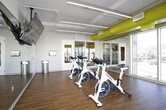 This single-level spin studio is designed to deliver an immersive cycling experience in a bright, well-appointed environment. The room is equipped with a row of professional Spinning-brand stationary bikes arranged to face a large, ceiling-mounted flat-screen television, allowing riders to follow guided workouts or entertainment during sessions. Full-length mirrored walls line one side of the studio, visually expanding the space and enabling riders to monitor their form throughout their workout.

A standout architectural feature is the expansive floor-to-ceiling windows that wrap around a corner of the room, flooding the interior with abundant natural light and offering open views of the surrounding urban neighborhood and skyline. The warm hardwood flooring provides both a polished aesthetic and a durable, easy-to-maintain surface suited to high-traffic fitness use.

Overhead, exposed ductwork painted in a bold yellow-green adds an industrial yet energetic design element that contributes to the studio's modern character. Linear LED lighting runs along the ceiling, ensuring consistent, even illumination throughout the space. The overall ambiance is clean, energizing, and purpose-built for group cycling classes, combining functional fitness design with contemporary architectural details.