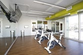 This single-level spin studio is designed to deliver an immersive cycling experience in a bright, well-appointed environment. The room is equipped with a row of professional Spinning-brand stationary bikes arranged to face a large, ceiling-mounted flat-screen television, allowing riders to follow guided workouts or entertainment during sessions. Full-length mirrored walls line one side of the studio, visually expanding the space and enabling riders to monitor their form throughout their workout.

A standout architectural feature is the expansive floor-to-ceiling windows that wrap around a corner of the room, flooding the interior with abundant natural light and offering open views of the surrounding urban neighborhood and skyline. The warm hardwood flooring provides both a polished aesthetic and a durable, easy-to-maintain surface suited to high-traffic fitness use.

Overhead, exposed ductwork painted in a bold yellow-green adds an industrial yet energetic design element that contributes to the studio's modern character. Linear LED lighting runs along the ceiling, ensuring consistent, even illumination throughout the space. The overall ambiance is clean, energizing, and purpose-built for group cycling classes, combining functional fitness design with contemporary architectural details.