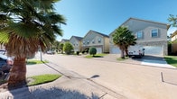This single-story home is situated within a tidy residential neighborhood characterized by stucco and stone-accented exteriors, attached garages, and mature palm trees lining the streets, lending a warm, suburban feel to the surroundings.

Inside, the home features rich, dark hardwood-style flooring that flows consistently through the main living areas, creating a cohesive and inviting atmosphere. The interior walls are finished in soft, neutral gray tones that complement the flooring and contribute to a clean, modern aesthetic. A standout architectural detail is the elegant arched doorway that connects the main living spaces, adding a classic touch to the otherwise contemporary design.

The living area offers a comfortable layout with ample natural light filtering in through blinds-covered windows, while wall art and decorative accents add personality to the space. An open work or desk area is also visible, suggesting flexible use of the interior space.

A hallway connects the rooms, with transitions between the hardwood flooring and carpeted areas noted in the bedroom zones. Wainscoting paneling in one room adds a refined architectural layer. Overall, the home presents a well-appointed, functional single-floor layout suitable for everyday living.