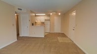 This single-floor unit features an open-concept layout that seamlessly connects the living area and kitchen, creating a cohesive and functional living space. The main living area is finished with wall-to-wall beige carpeting, lending a warm and neutral tone throughout. Warm beige walls complement the flooring, contributing to a consistent, understated aesthetic.

The kitchen is positioned centrally and defined by a raised peninsula countertop that serves as a natural divider between the cooking and living spaces. White cabinetry lines the kitchen walls, providing ample storage, and is paired with white appliances, including a full-sized refrigerator. A under-cabinet range hood is installed above the cooking area, and a sink with a window-side position adds practicality. Track lighting is mounted on the ceiling above the kitchen, providing focused illumination.

The unit includes multiple doors visible from the main living space, suggesting access to additional rooms, a closet, and the main entry. A louvered closet door is also visible, offering dedicated storage. The overall ambiance is clean, neutral, and move-in ready, suited for straightforward, comfortable living.