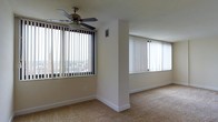 Unit 1205 is a single-floor residence that immediately impresses with its abundance of natural light. The open living area is lined with large windows dressed in vertical blinds, framing sweeping elevated views of the surrounding cityscape. Neutral-toned carpet flooring runs throughout, lending a warm and cohesive feel to the space. A ceiling fan is centrally positioned in the main living area, providing both functionality and a classic touch. The layout features a partial dividing wall that subtly defines areas within the open floor plan while maintaining an airy, connected atmosphere. Clean white walls and simple white baseboards keep the aesthetic understated and versatile, making the space easy to personalize. The high-rise positioning of the unit allows for bright, unobstructed light to fill the interior throughout the day. Overall, Unit 1205 offers a comfortable, light-filled environment with practical finishes and an open feel well-suited for everyday living.