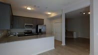 This two-story home showcases a thoughtfully designed open-concept main level, anchored by a well-appointed kitchen. The kitchen features dark espresso-toned cabinetry that provides generous storage, complemented by granite countertops with subtle veining. Stainless steel appliances, including a French door refrigerator with an ice and water dispenser, a range with electric cooktop, and an over-the-range microwave, give the kitchen a sleek, contemporary feel. A kitchen island with a white base extends the counter space and serves as a natural divider between the kitchen and the adjoining living area. Light hardwood-style flooring runs throughout the main level, creating a warm, cohesive aesthetic. A structural column and open sightlines connect the kitchen to the living space, where a staircase leads to the second floor. Closet doors near the base of the stairs suggest convenient main-level storage. Recessed and flush-mount ceiling lighting illuminate the space effectively. The overall design balances functionality and modern style across both levels.