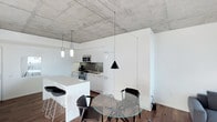 This single-floor space makes a striking first impression with its exposed concrete ceiling, which lends an industrial character balanced by crisp, clean white walls and cabinetry. The open-concept layout seamlessly connects the kitchen and dining areas, creating a cohesive and functional living environment. The kitchen is outfitted with sleek, handle-free white cabinetry, stainless steel appliances including a range and microwave, and a clean white countertop with an undermount sink. A white kitchen island with bar seating provides additional workspace and casual dining options. Pendant lighting in matte black and brushed metal tones descends from the concrete ceiling, adding a refined contrast to the minimalist palette. The dining area features a circular glass-top table paired with modern black chairs, contributing to the contemporary aesthetic. Rich, dark hardwood flooring runs throughout, grounding the space and adding warmth against the cooler tones of the concrete and white surfaces. The overall ambiance is one of urban sophistication — a thoughtfully designed space where industrial elements and modern finishes coexist in a clean, uncluttered setting.