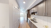 This efficient studio in Austin, TX offers a thoughtfully organized single-floor layout encompassing a kitchen, living area, and full bathroom. The galley-style kitchen is a standout feature, lined with taupe shaker-style cabinetry offering ample upper and lower storage, stainless steel appliances including a full-size refrigerator, dishwasher, and range, a single-basin stainless steel sink, light quartz countertops, and a classic white subway tile backsplash. An open display niche in the cabinetry adds a decorative touch. Warm-toned wood-look flooring runs seamlessly throughout the entire space, lending a cohesive and inviting feel. The living area is bright and clean, featuring white walls, a ceiling fan, and a window that draws in natural light. The open layout allows for flexible furniture arrangement. The bathroom is accessed via a paneled white door and continues the warm flooring, featuring clean white walls and a tiled shower area. A closet with paneled doors provides additional storage along the entry corridor. The overall design is modern and neutral, making for a versatile and functional living space in a vibrant Austin neighborhood.