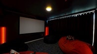 Located at 1000 Diamond Street in Philadelphia, PA, this purpose-built screening room delivers a fully immersive home theater experience within a compact, single-level layout. The room is finished entirely in deep black, from the walls and ceiling to the carpet-tiled flooring, creating a light-controlled environment optimized for viewing. A large, centrally mounted projection screen dominates one wall, flanked on either side by vivid red LED floor lamps that cast a dramatic, cinematic glow throughout the space. A ceiling-mounted projector ensures crisp, high-quality image projection. Thick floor-to-ceiling blackout curtains line one wall, further enhancing the darkened atmosphere while adding a polished, theatrical aesthetic. Seating is provided by multiple oversized red bean bag chairs arranged for comfortable, casual viewing. A wall-mounted mini-split HVAC unit ensures year-round climate control, and an additional projector or speaker component is mounted near the center of the room for enhanced audiovisual performance. A single entry door connects the theater to an adjacent space, partially visible through a frosted glass panel. Subtle track lighting along the ceiling perimeter adds ambient depth. This room is thoughtfully designed to provide a fully enclosed, distraction-free cinematic retreat.
