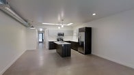 This contemporary single-floor space showcases a thoughtfully designed open-concept layout that emphasizes clean lines and modern finishes. The kitchen serves as the centerpiece of the space, featuring a striking contrast between dark espresso cabinetry and light upper cabinets. A central island with an integrated sink provides both functional workspace and a natural gathering point, illuminated by sleek pendant lighting suspended from above. Stainless steel appliances, including a French door refrigerator, a built-in microwave, and a range, lend a professional quality to the culinary space. Recessed lighting and a skylight fixture brighten the kitchen area, complementing the neutral white walls and smooth, polished concrete floors that extend seamlessly throughout the space. The open floor plan connects the kitchen to adjacent areas, with a long corridor visible in the background that hints at the depth and flow of the layout. An industrial-style pendant light anchors one end of the space, adding a subtle design accent. The overall ambiance is clean, minimalist, and contemporary, making for a versatile and visually cohesive environment.