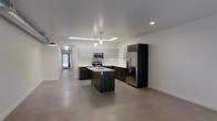 This contemporary single-floor space showcases a thoughtfully designed open-concept layout that emphasizes clean lines and modern finishes. The kitchen serves as the centerpiece of the space, featuring a striking contrast between dark espresso cabinetry and light upper cabinets. A central island with an integrated sink provides both functional workspace and a natural gathering point, illuminated by sleek pendant lighting suspended from above. Stainless steel appliances, including a French door refrigerator, a built-in microwave, and a range, lend a professional quality to the culinary space. Recessed lighting and a skylight fixture brighten the kitchen area, complementing the neutral white walls and smooth, polished concrete floors that extend seamlessly throughout the space. The open floor plan connects the kitchen to adjacent areas, with a long corridor visible in the background that hints at the depth and flow of the layout. An industrial-style pendant light anchors one end of the space, adding a subtle design accent. The overall ambiance is clean, minimalist, and contemporary, making for a versatile and visually cohesive environment.