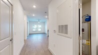 This single-floor unit is designed with a clean, contemporary aesthetic, characterized by crisp white walls, recessed lighting, and wood-look plank flooring that flows throughout the main living areas. The open layout connects the entryway corridor to a well-proportioned living area at the far end, where a ceiling fan and a pair of windows allow natural light to fill the space.

The kitchen is a functional highlight, featuring white shaker-style cabinetry with brushed nickel hardware, granite-look countertops, and a white subway tile backsplash. A full suite of stainless steel appliances includes a refrigerator, electric range, over-the-range microwave, and dishwasher.

The bedroom is carpeted for comfort, outfitted with a ceiling fan, and includes two windows offering natural light. A spacious walk-in closet with wire shelving provides ample storage. The bathroom features a white vanity with granite countertop, a large wall mirror, wood-look flooring, and a shower-tub combination.

A dedicated laundry area houses a full-size Whirlpool top-load washer and dryer with wire shelving above. The HVAC system includes a thermostat and ventilation panel near the entry. Overall, the unit presents a bright, move-in-ready environment with practical, modern finishes throughout.