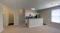 Located at 2600 Harvest Creek Pl in Cary, North Carolina, this single-floor residence offers a thoughtfully designed open-concept layout that seamlessly connects the kitchen and living areas. The kitchen is a standout feature, showcasing white cabinetry, a mosaic tile backsplash, and a full suite of stainless steel appliances, including a refrigerator, microwave, and range. A central island with dark countertops provides additional prep space and serves as a natural dividing point between the kitchen and the carpeted living area. Flush-mount ceiling lighting illuminates both zones evenly, contributing to a clean and functional ambiance. The neutral color palette — soft beige walls, white trim, and warm carpet — creates a cohesive, move-in-ready aesthetic. A hallway visible from the main living space leads to additional rooms, suggesting a practical and well-organized floor plan. The space is designed with everyday comfort and convenience in mind, making it an excellent example of modern, low-maintenance living in the heart of Cary, NC.