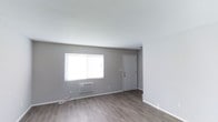 This two-floor space showcases a straightforward, functional design characterized by a neutral grey and white color palette that creates a clean, modern aesthetic. The main living area, visible in the provided image, features smooth white ceilings and grey walls accented by crisp white baseboards, contributing to a bright and open feel. Dark hardwood-style flooring runs throughout, offering durability and a warm contrast to the lighter wall tones. A window along the exterior wall allows natural light to fill the room, while a wall-mounted air conditioning unit provides climate control. A paneled white interior door adds a classic architectural detail to the space. The overall layout appears straightforward and open, offering flexibility for various furniture arrangements. The minimalist finish and neutral tones provide a versatile backdrop, making the space adaptable to a range of interior styles. The two-floor layout suggests additional living or functional areas beyond what is immediately visible, potentially expanding the overall usable space considerably.