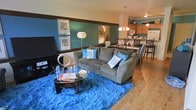 This 3-floor space showcases a thoughtfully designed open-concept main living area where the living room and kitchen flow seamlessly into one another. The living room is anchored by warm hardwood flooring and features a rich teal accent wall adorned with framed artwork, creating a bold and inviting focal point. A wide, grey upholstered sofa dressed with blue decorative pillows sits atop a vibrant blue shag area rug, adding a layer of texture and color contrast to the space. A glass-top coffee table and a wall-mounted flat-screen television complete the living arrangement.

The open layout transitions naturally into a dining area, where a dark-toned dining table and bar-height seating define the space. Beyond the dining area, the kitchen is equipped with warm wood cabinetry, a white refrigerator, and pendant lighting that adds warmth overhead. A dark horizontal trim band runs along the upper wall throughout the space, tying together the color scheme.

The multi-floor layout offers additional living space across its three levels, providing versatility and room to accommodate a range of needs. The overall ambiance is lively and contemporary, driven by a cohesive blue and brown color palette.