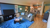This 3-floor space showcases a thoughtfully designed open-concept main living area where the living room and kitchen flow seamlessly into one another. The living room is anchored by warm hardwood flooring and features a rich teal accent wall adorned with framed artwork, creating a bold and inviting focal point. A wide, grey upholstered sofa dressed with blue decorative pillows sits atop a vibrant blue shag area rug, adding a layer of texture and color contrast to the space. A glass-top coffee table and a wall-mounted flat-screen television complete the living arrangement.

The open layout transitions naturally into a dining area, where a dark-toned dining table and bar-height seating define the space. Beyond the dining area, the kitchen is equipped with warm wood cabinetry, a white refrigerator, and pendant lighting that adds warmth overhead. A dark horizontal trim band runs along the upper wall throughout the space, tying together the color scheme.

The multi-floor layout offers additional living space across its three levels, providing versatility and room to accommodate a range of needs. The overall ambiance is lively and contemporary, driven by a cohesive blue and brown color palette.