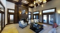 This distinguished space makes an immediate impression with its dramatic soaring ceilings adorned with rich, dark-stained coffered woodwork that adds depth and architectural character throughout. A commanding floor-to-ceiling fireplace surround, crafted in warm dark wood paneling, serves as the room's focal centerpiece and is accented by a striking starburst mirror that draws the eye upward. An elegant multi-arm chandelier with candle-style lighting hangs overhead, complementing the recessed lighting to create a warm, layered glow throughout the space. Tall, multi-pane windows and glass-paneled doors flood the interior with natural light while offering views of the outdoor surroundings, seamlessly connecting the interior to the exterior. The seating area is thoughtfully arranged with soft upholstered armchairs and a neutral-toned sofa, centered around a bold dark wood coffee table with decorative cross-base detailing. Fresh white floral arrangements add a crisp, inviting touch to the overall aesthetic. The flooring features light-toned tile, partially layered with a textured area rug, contributing to the space's refined yet comfortable character. Overall, this space exudes a blend of classic elegance and contemporary sophistication.