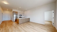 This single-floor unit showcases a clean, contemporary design characterized by crisp white walls, light hardwood flooring, and a cohesive neutral palette throughout. The open-concept living and kitchen area forms the heart of the space, where a central island with a white quartz countertop anchors the kitchen. Two-tone cabinetry—combining warm walnut-toned lower cabinets with lighter upper cabinets—complements a full suite of stainless steel appliances, including a refrigerator, gas range, built-in microwave, and dishwasher. Recessed lighting illuminates the kitchen and living area, enhancing the sense of openness.

The hallway features a built-in recessed shelf, offering a practical yet streamlined storage solution, alongside dark wood accent cabinetry that adds visual depth. The bedrooms are well-proportioned, with one room benefiting from large floor-to-ceiling windows that admit generous natural light and frame views of surrounding greenery and neighboring rooftops.

The bathroom maintains the same modern aesthetic, featuring warm wood vanity cabinetry, a white countertop with an undermount sink, and a glass-enclosed shower. Light hardwood flooring runs continuously throughout all areas, reinforcing the unit's cohesive and contemporary character.