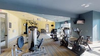 This fitness center is a thoughtfully designed, single-floor facility offering a comprehensive workout environment for users of all fitness levels. The space features a warm yellow and steel-blue color palette, complemented by striped carpeted flooring that adds a contemporary aesthetic throughout.

Large floor-to-ceiling mirrors line one wall, visually expanding the space and allowing users to monitor their form during exercise. Natural light filters in through windows and a glass-paneled exit door, brightening the interior alongside recessed ceiling lighting and ceiling fans that promote air circulation.

The cardio zone is well-appointed with treadmills, stationary bikes — including Expresso interactive cycling units — and elliptical machines, several equipped with personal screens for entertainment or guided workouts. A dedicated strength training area houses a Hoist multi-station cable machine along with a full dumbbell rack and adjustable weight bench to support a range of resistance training routines.

A functional training section adds versatility, featuring an Iron Cage multi-use rig, a heavy punching bag, a mini trampoline, battle ropes, and resistance bands. Wall-mounted televisions are positioned throughout for added convenience. The overall ambiance is clean, open, and energizing, making it a well-rounded facility suited for cardio, strength, and functional fitness training.