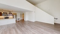 This two-story space is designed with a clean, contemporary aesthetic that emphasizes light and openness. The main living area makes an immediate impression with its double-height ceilings and expansive floor-to-ceiling windows wrapping two walls, flooding the interior with natural light and offering views of the surrounding urban streetscape. Light wood-tone flooring runs throughout, reinforcing the bright, airy atmosphere. A staircase leads to the upper level, where a mezzanine overlooks the living space below, adding architectural depth and a sense of vertical scale.

The kitchen is thoughtfully designed with a quartz waterfall-edge peninsula that doubles as a prep and dining surface. Light wood cabinetry on the lower level is paired with sleek high-gloss upper cabinets in a muted blue-grey tone. Stainless steel appliances, including a gas range, built-in microwave, and refrigerator, are complemented by track and pendant lighting overhead.

The bedroom features exposed concrete ceiling details, a window, and two generously sized closets with wood rod-and-shelf systems. A dedicated walk-in closet with double hanging rods and shelving provides additional storage. The bathroom includes a wood-grain vanity with a quartz countertop, a large mirror, and a soaking tub with white tile surround.