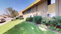 This single-story rental office presents a welcoming and professionally maintained exterior designed for easy accessibility and curb appeal. The entrance is fronted by a covered walkway with classic brick-paved flooring, illuminated by ceiling-mounted light fixtures that provide warm, consistent lighting throughout the day and evening. A bold "Rental Office" sign marks the entry clearly, while large glass storefront windows and a glass door allow natural light to flood the space and offer a transparent view into the interior, which features decorative plants and a vending machine visible from outside.

The building exterior is finished in a warm yellow and tan stucco combination, lending it a distinct and approachable character. A wrought iron railing lines the covered patio area, offering a slight separation from the surrounding landscape. The grounds are thoughtfully landscaped with trimmed shrubs, flowering plants, and a well-kept lawn that wraps around the building, enhancing the overall setting. Mature trees provide natural shade and a sense of established greenery throughout the property. The surrounding area includes ample parking and neighboring residential buildings, all set beneath open, sunny skies that reflect a pleasant Southern California-style environment.