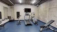 This fitness center offers a well-equipped workout space designed to accommodate a range of exercise needs. The space is characterized by its industrial aesthetic, featuring exposed concrete walls, visible ceiling ductwork, and raw structural beams that lend the room a distinctive, urban character. The flooring throughout is a durable blue rubberized surface, well-suited for high-impact activity and easy maintenance.

The facility is divided into two functional zones. One area is dedicated to cardio and aerobic training, housing a treadmill, elliptical machine, stationary spin bike, and a stability ball, all positioned beneath a wall-mounted flat-screen television — ideal for staying entertained during workouts. The second area focuses on strength training, featuring a multi-function cable machine, an adjustable weight bench, and a fully stocked dumbbell rack with a wall mirror for form monitoring. A wall-mounted sanitizing station and waste bin are conveniently accessible, supporting cleanliness throughout the space. Overhead linear lighting and a ceiling-mounted HVAC unit ensure the environment remains well-lit and climate-controlled during use. A glass-panel door connects to an adjacent area, adding accessibility and openness to the layout.