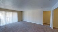 This single-floor residential space offers a clean, straightforward layout designed for comfortable everyday living. The main living area is finished with wall-to-wall carpeting in a neutral tone, providing a soft and warm underfoot feel throughout. The walls are painted in a light, creamy beige palette that creates a bright and welcoming atmosphere, complemented by crisp white baseboards that add a clean finish to the room.

A large window dressed with vertical blinds allows for adjustable natural light, making the space feel open and airy. The smooth, flat ceiling enhances the sense of openness and keeps the aesthetic simple and uncluttered. A smoke detector is visibly installed, reflecting attention to safety.

An adjacent room, accented with a warm golden-yellow wall tone, is accessible from the main living area, offering a visual contrast and a sense of defined spatial flow between rooms. The overall design of the space is practical and neutral, making it a versatile canvas well-suited for a variety of furnishing styles and personal tastes. The layout is efficient, with clear sightlines between areas that contribute to a cohesive and functional living environment.