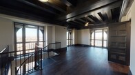 This sophisticated two-floor space blends bold architectural character with modern finishes throughout. The upper level serves as a dramatic centerpiece, showcasing exposed dark wood beam ceilings, rich dark hardwood flooring, and an expansive open layout flooded with natural light from large, arched-accent windows on multiple sides. Sweeping panoramic views of the surrounding cityscape and landscape are visible from nearly every angle, lending the space an elevated, airy quality. A sleek iron staircase with black metal railings connects both levels with understated elegance.

The kitchen is well-appointed with white cabinetry, subway tile backsplash, stainless steel appliances including a full-size refrigerator, range, and microwave, along with a generous island topped in white quartz — ideal for both cooking and casual gatherings. The coffered, beamed ceiling continues here, adding depth and visual interest overhead.

The bedrooms are bright and functional, each featuring dark hardwood floors, ceiling fans, and large windows offering city views. The bathrooms are cleanly finished — one features a white vanity with a sliding glass shower enclosure, while another showcases striking marble-effect wall panels and a quartz countertop vanity. A stacked in-unit washer and dryer provides added everyday convenience.