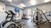 This fitness center in Laurel, Maryland offers a comprehensive workout facility thoughtfully arranged across a single open floor. The space is finished with durable rubber flooring throughout, providing a safe and practical surface for a wide range of exercises. Soft gray walls and bright overhead fluorescent lighting create a clean, energizing environment conducive to focused training.

The cardio area is well-stocked with a treadmill, elliptical cross-trainer, and a step machine, all positioned toward the front of the space. A large sliding glass door with vertical blinds allows natural light to filter in and offers a view of the surrounding greenery, adding a refreshing outdoor connection during workouts. A wall-mounted flat-screen television provides entertainment while exercising.

Strength training equipment includes a full multi-station Hoist cable machine suitable for a variety of resistance exercises, complemented by a tiered dumbbell rack stocked with a complete set of free weights. A body weight scale is also available for tracking fitness progress. Instructional fitness charts and equipment guides are posted on the walls for user reference.

An adjacent office area with vaulted ceilings, glass-paneled doors, carpeted flooring, and a computer workstation appears to serve as an administrative or leasing office connected to the fitness facility.