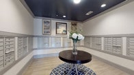 This thoughtfully designed mail room serves as both a functional and visually appealing space within a residential building. The room is defined by its striking dark navy ceiling, which contrasts elegantly with the light gray walls, creating a sophisticated and polished atmosphere. Recessed lighting is evenly distributed across the ceiling, ensuring the space is well-lit throughout.

The walls are lined with extensive banks of individual mailboxes finished in a coordinating gray tone, providing ample mail storage for residents. The mailboxes are arranged in neat, uniform rows across multiple walls, maximizing capacity while maintaining a clean and organized appearance. A wainscoting-style horizontal molding rail runs along the walls, adding a layer of architectural detail.

Above the mailbox rows, three framed art pieces are displayed, lending a gallery-like touch to the utilitarian space. Light wood-look flooring anchors the room warmly, while a patterned area rug in a classic Moroccan tile design adds visual interest underfoot. At the center of the room, a round dark wood table serves as a focal point, topped with an elegant glass vase of fresh white floral arrangements, infusing the space with a welcoming and upscale feel.