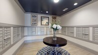 This thoughtfully designed mail room serves as both a functional and visually appealing space within a residential building. The room is defined by its striking dark navy ceiling, which contrasts elegantly with the light gray walls, creating a sophisticated and polished atmosphere. Recessed lighting is evenly distributed across the ceiling, ensuring the space is well-lit throughout.

The walls are lined with extensive banks of individual mailboxes finished in a coordinating gray tone, providing ample mail storage for residents. The mailboxes are arranged in neat, uniform rows across multiple walls, maximizing capacity while maintaining a clean and organized appearance. A wainscoting-style horizontal molding rail runs along the walls, adding a layer of architectural detail.

Above the mailbox rows, three framed art pieces are displayed, lending a gallery-like touch to the utilitarian space. Light wood-look flooring anchors the room warmly, while a patterned area rug in a classic Moroccan tile design adds visual interest underfoot. At the center of the room, a round dark wood table serves as a focal point, topped with an elegant glass vase of fresh white floral arrangements, infusing the space with a welcoming and upscale feel.