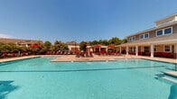 Located at 1000 Broadstone Way in Apex, NC, this outdoor amenity space is designed to offer a resort-style recreational experience. The centerpiece is a generously sized swimming pool with crystal-clear blue water, accessible via wide entry steps and surrounded by an expansive paver deck. Red lounge chairs are arranged throughout the deck area, providing ample space for sunbathing and relaxation under the open North Carolina sky.

Adjacent to the pool, a covered pergola structure serves as a shaded outdoor living and entertainment hub. Beneath it, a built-in brick bar counter with a granite-style top offers a functional gathering space, complemented by red cushioned seating. A ceiling fan overhead ensures comfort during warm days. A dedicated grilling station with a stainless steel grill is positioned nearby, making the area well-suited for outdoor cooking and social gatherings.

The clubhouse building anchors the space architecturally, featuring classic craftsman-style detailing with gray lap siding, brick wainscoting, red accent doors, and white-trimmed windows. A covered front porch equipped with ceiling fans and additional red lounge furniture provides a shaded retreat directly overlooking the pool. The entire area is enclosed by decorative iron fencing, with mature trees lining the perimeter, offering both privacy and a lush, green backdrop.
