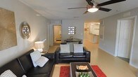 This single-floor residence showcases an open-concept design that integrates the living, dining, and kitchen areas into one cohesive space. The living room serves as the central gathering point, furnished with a sleek black leather sectional sofa accented by patterned throw pillows. A bold red area rug and a dark wood coffee table add contrast and visual interest to the neutral-toned flooring throughout. A ceiling fan with an integrated light fixture provides both comfort and illumination overhead.

Just beyond the living area, a compact dining space features a dark table set against a wall adorned with a decorative glass block window, allowing diffused natural light to filter through. The open passageway leads directly into the kitchen, which is visible in the background and features light cabinetry and a raised breakfast bar, suggesting additional seating and workspace.

A doorway off the living area provides access to a bathroom, which is partially visible and appears clean and functional. The overall palette is neutral and contemporary, with light walls and flooring creating a bright, airy ambiance throughout the single-level layout.