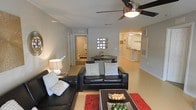 This single-floor residence showcases an open-concept design that integrates the living, dining, and kitchen areas into one cohesive space. The living room serves as the central gathering point, furnished with a sleek black leather sectional sofa accented by patterned throw pillows. A bold red area rug and a dark wood coffee table add contrast and visual interest to the neutral-toned flooring throughout. A ceiling fan with an integrated light fixture provides both comfort and illumination overhead.

Just beyond the living area, a compact dining space features a dark table set against a wall adorned with a decorative glass block window, allowing diffused natural light to filter through. The open passageway leads directly into the kitchen, which is visible in the background and features light cabinetry and a raised breakfast bar, suggesting additional seating and workspace.

A doorway off the living area provides access to a bathroom, which is partially visible and appears clean and functional. The overall palette is neutral and contemporary, with light walls and flooring creating a bright, airy ambiance throughout the single-level layout.