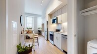 This single-floor studio is designed to maximize functionality within an open-concept layout. Impressively tall ceilings lend a sense of spaciousness throughout, while light hardwood flooring flows seamlessly from room to room. A large window at the far end draws in abundant natural light and offers views of the surrounding streetscape.

The galley-style kitchen is well-appointed with stainless steel appliances, including a refrigerator, electric range, built-in microwave, and dishwasher. Light wood cabinetry extends floor to ceiling, providing generous storage, while a white subway tile backsplash and light stone countertops complete the clean, contemporary aesthetic. Under-cabinet lighting enhances visibility and adds a polished touch.

The main living area integrates a built-in Murphy bed with an integrated patterned headboard panel and flanking shelving units, offering a practical sleeping solution without sacrificing living space. A built-in desk nook provides a dedicated workspace. The open floor plan comfortably accommodates a seating area alongside the dining and kitchen zones.

The bathroom features light tile flooring, a single-sink vanity with storage cabinetry, a toilet, and a walk-in shower with a curtained enclosure. The overall ambiance is bright, modern, and efficiently organized.
