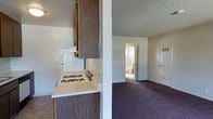 This single-floor space offers a practical and straightforward layout designed for comfortable everyday living. The interior is characterized by a textured popcorn ceiling that runs consistently throughout, lending a cohesive feel to the space.

The kitchen is partially open to the living area, separated by a half-wall partition that creates a sense of definition while maintaining an open flow between the two spaces. It features dark wood cabinetry for ample storage, light-colored countertops, and a gas range with a white finish. The kitchen floor is finished with tile, providing a durable and easy-to-clean surface.

The adjoining living area is carpeted in a deep plum-toned carpet, adding warmth and texture underfoot. The neutral, light-toned walls keep the space feeling bright and airy, and multiple electrical outlets are conveniently positioned throughout.

A hallway visible from the kitchen leads to the bathroom, which appears to include standard fixtures and benefits from a window that brings in natural light. An entry door is also visible from this vantage point, offering a clear and accessible path through the space.

Overall, the layout is efficient and functional, making good use of the available space with clear transitions between the kitchen, living, and bathroom areas.