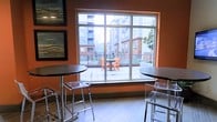 This single-floor space features a warm, inviting interior defined by rich terracotta-orange walls that create an energetic yet comfortable atmosphere. Large picture windows dominate one wall, flooding the interior with natural light and offering clear views of an adjacent outdoor patio and surrounding multi-story buildings. The seamless visual connection between indoors and outdoors enhances the sense of openness throughout the space.

Inside, high-top bistro tables paired with modern acrylic ghost chairs provide casual seating well-suited for socializing or working. The light wood-toned flooring adds a clean, contemporary contrast to the bold wall color. Framed artwork adorns the walls, contributing to a polished, thoughtfully curated aesthetic.

The adjoining outdoor terrace, visible through the expansive windows, features matching orange patio furniture, reinforcing a cohesive design language between the interior and exterior areas. Decorative planters and railings define the patio boundaries, creating an attractive alfresco seating option. Overall, the space balances a lively, modern design with functional, comfortable amenities suited to a community or hospitality setting.