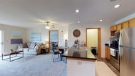 This single-floor space is thoughtfully designed around an open-concept layout that brings the kitchen, dining, and living areas together in one cohesive, flowing environment. The kitchen anchors the space with a dark-topped island/peninsula that doubles as a breakfast bar, featuring a double sink and ample counter space. Warm wood-tone door and window trim accents are carried consistently throughout, adding a welcoming character to the interior. Recessed lighting is installed throughout the ceiling, complemented by a ceiling fan with light fixture in the living area, ensuring the space is well-lit and comfortable year-round. The living area features plush carpeting and is furnished to accommodate relaxed seating, with coastal-themed wall art adding a light, airy feel. A small dining nook sits centrally within the open area, offering an intimate space for meals. Wood-look flooring in the kitchen and entryway zones provides a practical contrast to the soft carpet elsewhere. A doorway from the main living area leads directly into a bedroom, visible in the background, which appears bright and comfortably sized. An additional interior door suggests further utility or storage space. Overall, the layout is efficient and well-connected, making it a functional and inviting single-level living environment.