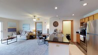 This single-floor space is thoughtfully designed around an open-concept layout that brings the kitchen, dining, and living areas together in one cohesive, flowing environment. The kitchen anchors the space with a dark-topped island/peninsula that doubles as a breakfast bar, featuring a double sink and ample counter space. Warm wood-tone door and window trim accents are carried consistently throughout, adding a welcoming character to the interior. Recessed lighting is installed throughout the ceiling, complemented by a ceiling fan with light fixture in the living area, ensuring the space is well-lit and comfortable year-round. The living area features plush carpeting and is furnished to accommodate relaxed seating, with coastal-themed wall art adding a light, airy feel. A small dining nook sits centrally within the open area, offering an intimate space for meals. Wood-look flooring in the kitchen and entryway zones provides a practical contrast to the soft carpet elsewhere. A doorway from the main living area leads directly into a bedroom, visible in the background, which appears bright and comfortably sized. An additional interior door suggests further utility or storage space. Overall, the layout is efficient and well-connected, making it a functional and inviting single-level living environment.