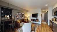 This single-floor residence is designed with a cohesive, contemporary aesthetic that balances style and functionality. The open-concept living area serves as the heart of the home, featuring warm hardwood-style flooring, neutral gray walls, and a wall-mounted flat-screen TV above a floating walnut media console. Seating is arranged around a patterned area rug, with a leather sofa accented by mustard and olive throw pillows lending a rich, inviting tone. Framed architectural artwork and a bar-height workspace with industrial stools add character to the space.

The kitchen is outfitted with dark espresso cabinetry extending to the ceiling, light countertops, and a full suite of black appliances, including a range, microwave, dishwasher, and refrigerator. A sliding glass door adjacent to the kitchen provides access to an outdoor balcony area, introducing natural light.

The bedroom features a dark accent wall with a display ledge for framed artwork, creating a focused, restful atmosphere. Hardwood-style flooring continues throughout.

The bathroom includes a full soaking tub with shower, accompanied by a stacked in-unit washer and dryer conveniently located nearby. Recessed lighting and clean finishes are consistent throughout the residence.