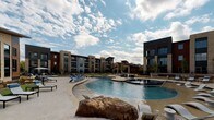 Located along Texas Park Road in Grand Prairie, Texas, this single-story amenity space is part of a contemporary multi-building residential community. The exterior architecture showcases a striking combination of natural limestone and brick cladding, paired with dark steel structural elements and warm wood-paneled ceilings that create a refined yet welcoming aesthetic.

The covered entrance features bold steel columns framing glass double doors, accented by a branded welcome mat and a sleek bench for guests. The overhang extends generously, providing shade over a smooth concrete walkway bordered by native landscaping with ornamental grasses, low shrubs, and gravel beds that reflect a Texas-inspired xeriscape design.

A black powder-coated mesh fence with a gated entry defines the perimeter of a dedicated outdoor area, offering access to manicured garden beds and connecting pathways throughout the community. Beyond the gate, a resort-style pool serves as a central outdoor focal point, featuring a freeform design with an adjacent spa, surrounding lounge chairs, and wide sun decks. The pool area is framed by the multi-story residential buildings, creating an enclosed, community-centered atmosphere under expansive Texas skies.