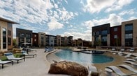 Located along Texas Park Road in Grand Prairie, Texas, this single-story amenity space is part of a contemporary multi-building residential community. The exterior architecture showcases a striking combination of natural limestone and brick cladding, paired with dark steel structural elements and warm wood-paneled ceilings that create a refined yet welcoming aesthetic.

The covered entrance features bold steel columns framing glass double doors, accented by a branded welcome mat and a sleek bench for guests. The overhang extends generously, providing shade over a smooth concrete walkway bordered by native landscaping with ornamental grasses, low shrubs, and gravel beds that reflect a Texas-inspired xeriscape design.

A black powder-coated mesh fence with a gated entry defines the perimeter of a dedicated outdoor area, offering access to manicured garden beds and connecting pathways throughout the community. Beyond the gate, a resort-style pool serves as a central outdoor focal point, featuring a freeform design with an adjacent spa, surrounding lounge chairs, and wide sun decks. The pool area is framed by the multi-story residential buildings, creating an enclosed, community-centered atmosphere under expansive Texas skies.