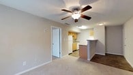 Tally Square is a well-designed single-floor residence that offers a practical and comfortable layout. The living area is spacious and inviting, finished with soft wall-to-wall carpeting and neutral tones that create a warm, welcoming atmosphere. A ceiling fan with a built-in light fixture is centrally mounted, providing both airflow and ambient lighting. The living space flows seamlessly into the kitchen via a peninsula-style breakfast bar, making the layout ideal for both everyday living and entertaining. The kitchen is efficiently organized with warm wood-toned cabinetry, granite-style countertops, and white appliances including a range, dishwasher, and refrigerator. Wood-look flooring in the kitchen adds a durable, easy-to-clean surface that contrasts nicely with the carpeted living area. The bedroom is generously sized, carpeted for comfort, and features a window with vertical blinds that allows natural light to filter in. It includes direct access to a private bathroom, which is outfitted with a wood-toned vanity, granite-style countertop, a large mirror, and wood-look flooring. Throughout the space, textured ceilings, clean white trim, and a consistent neutral color palette contribute to a cohesive and tidy aesthetic.