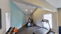 This dedicated fitness space is thoughtfully designed to support a full range of workout activities within a single-floor layout. The space is divided into two connected zones, each serving a distinct fitness purpose. The first zone features vaulted ceilings with recessed lighting and is painted in a calming light blue, providing an energizing atmosphere for cardio workouts. It houses an elliptical trainer, a treadmill, and additional cardio equipment, all set on durable dark carpet flooring. A window with blinds allows natural light to filter into the area.

The second zone transitions into a taller-ceilinged strength training area with neutral gray walls and continued dark carpet flooring. This section is equipped with a multi-function cable weight machine paired with an adjustable workout bench, a dumbbell rack with multiple weight sets, and a pull-up bar. A posted fitness rules board and workout chart add a structured, gym-like feel. Recessed ceiling lights provide consistent illumination throughout. A full-light exterior door in this zone opens directly to an outdoor area, offering convenient ventilation and easy access. Built-in open shelving provides practical storage within the space.