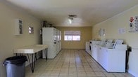 This single-floor communal laundry facility is designed for practicality and efficiency. The room is laid out with rows of full-sized washing machines and dryers lining both sides, providing ample capacity for multiple users at once. The machines are arranged in a clean, organized fashion, maximizing the usable floor space in the center of the room. A sturdy folding table is positioned along one wall, offering a convenient surface for sorting and folding laundry. The flooring consists of light-colored ceramic tiles that are easy to maintain and keep clean. Natural light enters through a centrally located window fitted with blinds, brightening the interior and creating a more pleasant environment. The walls are finished in a neutral yellow tone, contributing to the room's functional yet welcoming atmosphere. Overhead, a ceiling-mounted light fixture provides consistent illumination throughout the space. Ventilation is supported by a ceiling vent, ensuring proper airflow. Informational signage is posted on the walls, offering users guidance on machine operation and facility rules. Overall, the space is thoughtfully organized to serve as a convenient and fully functional shared laundry facility.