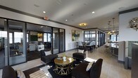 This single-floor space is designed as a multifunctional communal area with a refined, contemporary aesthetic. The open layout is anchored by a central lounge zone featuring dark upholstered armchairs and a bold black-and-white geometric area rug. A gold-finished round coffee table serves as a stylish focal point, accenting the space's black, white, and gold color scheme.

Glass-partitioned rooms line the perimeter, offering semi-private areas visible through sleek black-framed sliding glass doors. One enclosed room is outfitted with a chalkboard wall and work surfaces, suggesting a creative or collaborative workspace. Another glass-enclosed area appears to function as a private meeting or dining room, furnished with high-back chairs around a dark table.

Recessed lighting throughout the ceiling provides consistent, even illumination, while a decorative gold chandelier adds warmth above the dining area. Crown molding with dark trim detailing lends a polished architectural finish to the space. Natural light filters in through windows along the far wall, brightening the overall atmosphere. The space balances professional functionality with residential comfort.