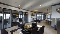 This single-floor space is designed as a multifunctional communal area with a refined, contemporary aesthetic. The open layout is anchored by a central lounge zone featuring dark upholstered armchairs and a bold black-and-white geometric area rug. A gold-finished round coffee table serves as a stylish focal point, accenting the space's black, white, and gold color scheme.

Glass-partitioned rooms line the perimeter, offering semi-private areas visible through sleek black-framed sliding glass doors. One enclosed room is outfitted with a chalkboard wall and work surfaces, suggesting a creative or collaborative workspace. Another glass-enclosed area appears to function as a private meeting or dining room, furnished with high-back chairs around a dark table.

Recessed lighting throughout the ceiling provides consistent, even illumination, while a decorative gold chandelier adds warmth above the dining area. Crown molding with dark trim detailing lends a polished architectural finish to the space. Natural light filters in through windows along the far wall, brightening the overall atmosphere. The space balances professional functionality with residential comfort.